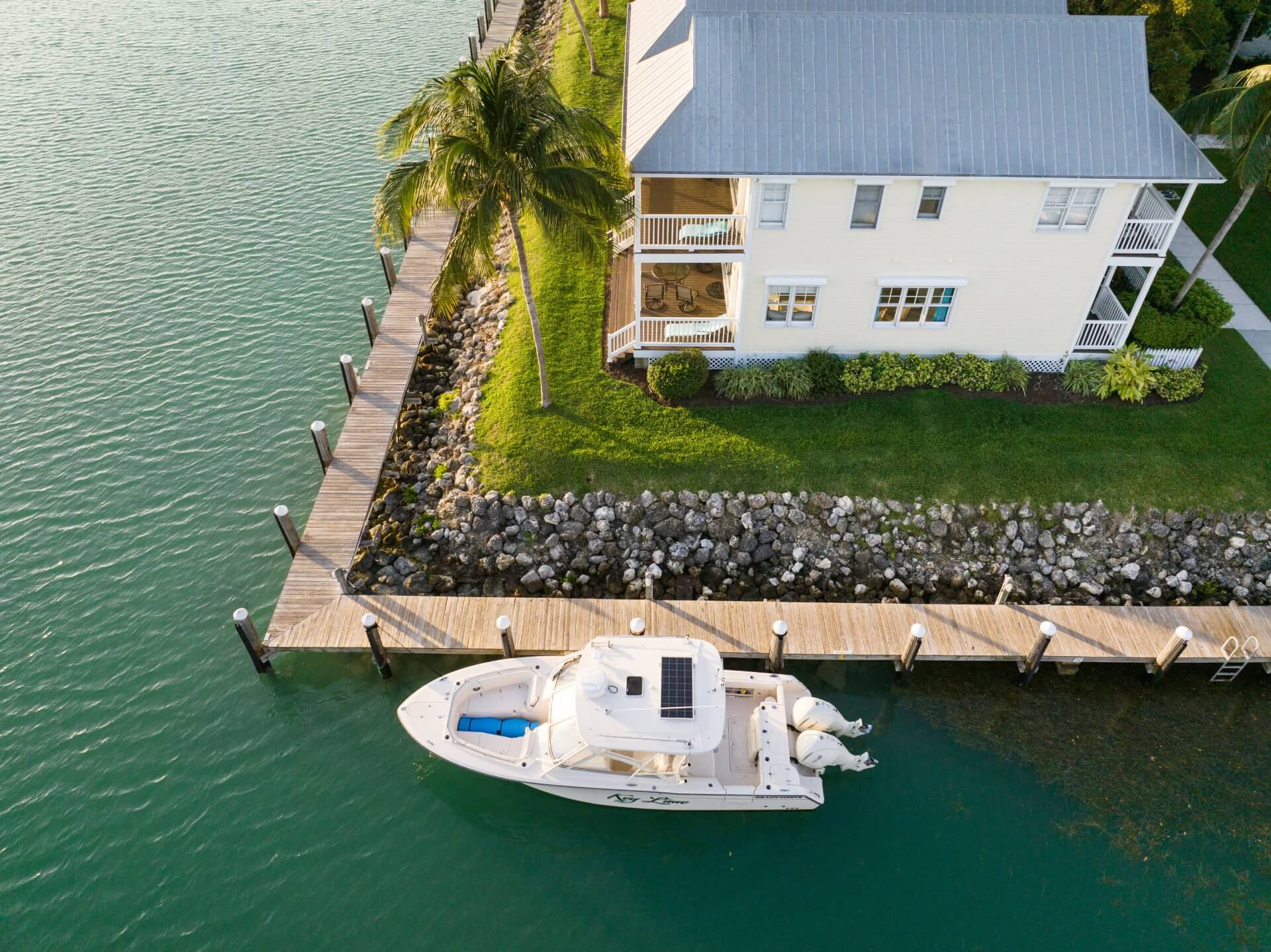 Aerial view of waterfront home with private dock, palm trees, and a boat moored along calm turquoise water.