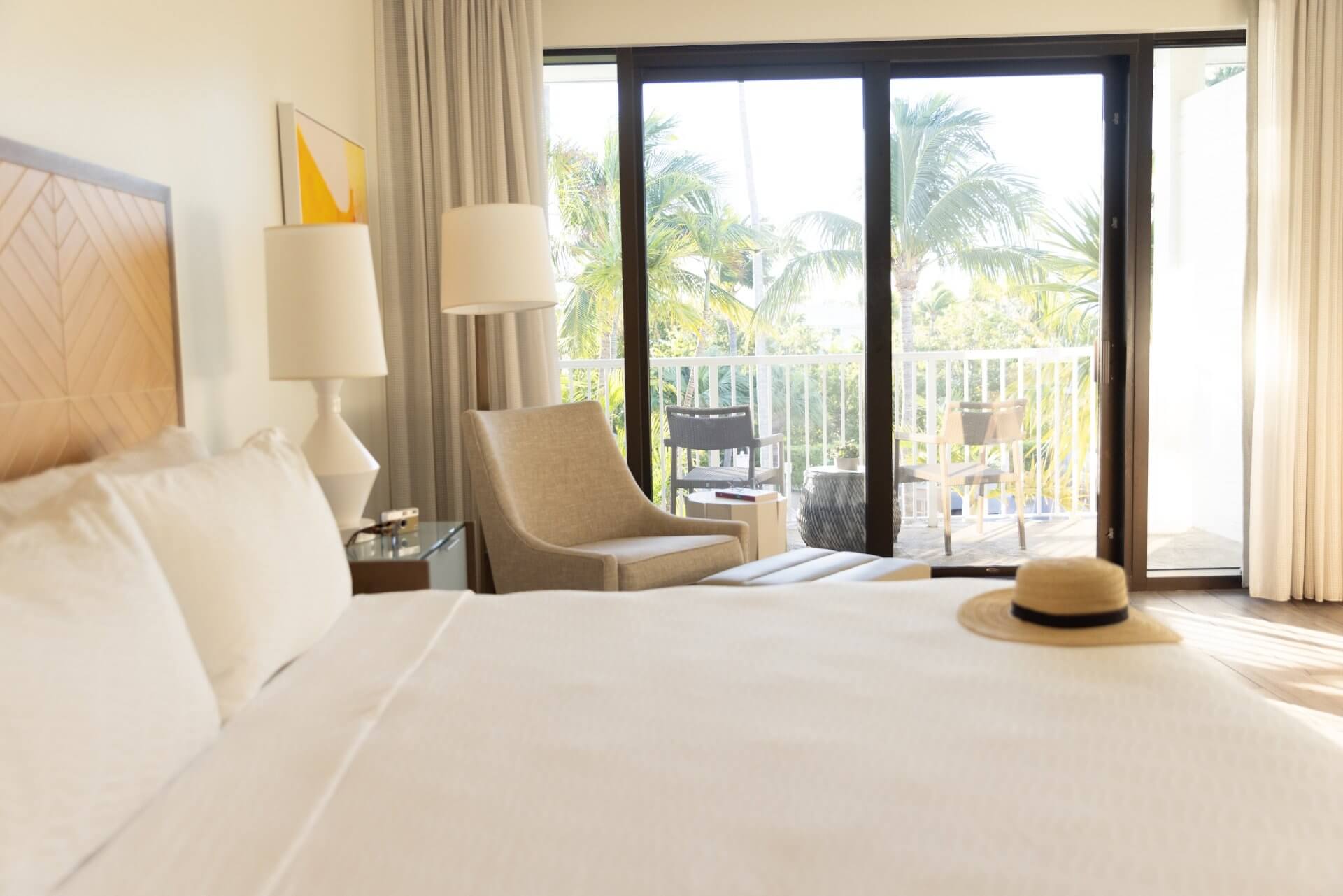 Bright hotel room with a king bed, lounge chair, and sliding doors opening to a balcony with palm tree views.