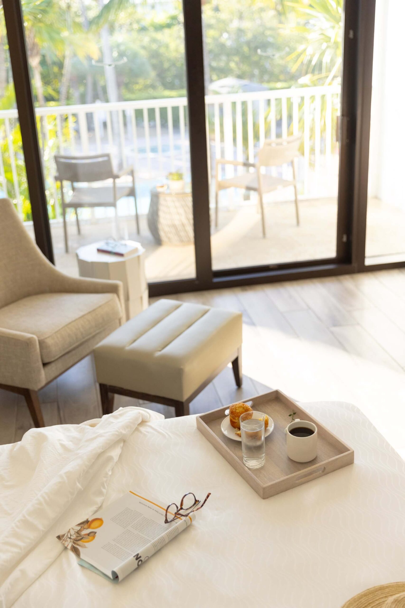 Sunlit bedroom with coffee tray on the bed, reading glasses and magazine, and balcony seating visible through sliding doors.