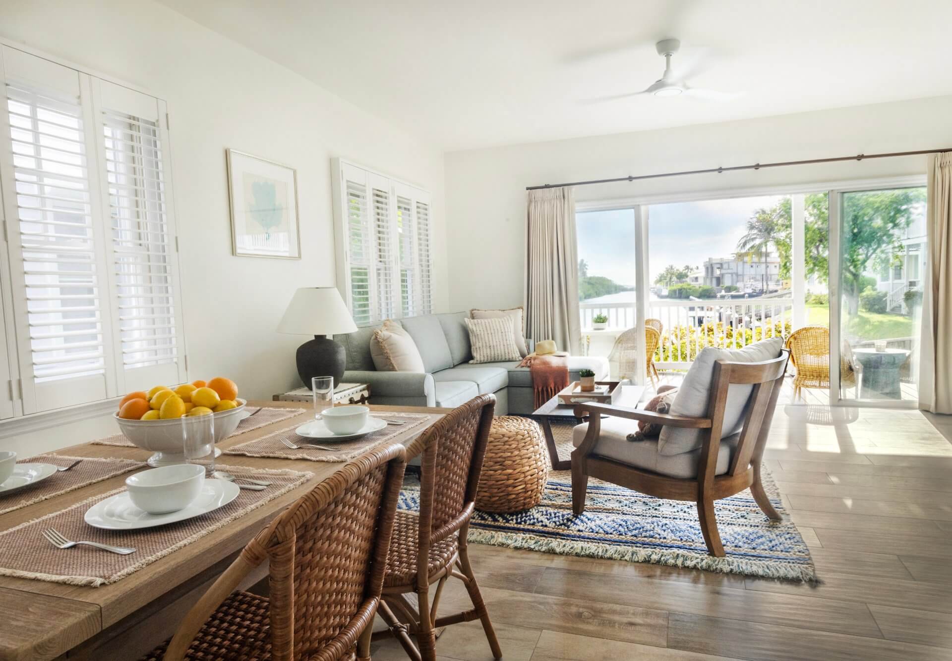 Bright coastal living and dining area with wicker accents, neutral tones, and sliding doors opening to a sunny balcony.