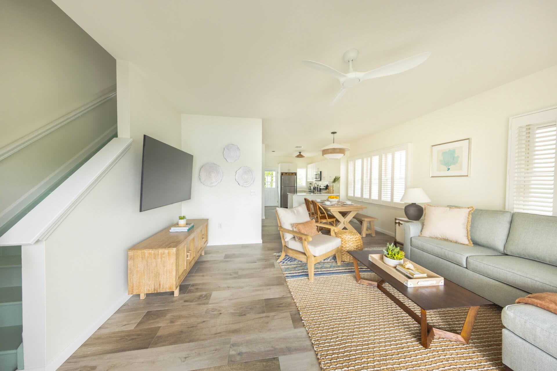 Bright coastal living room with gray sofa, wood furniture, woven rug, and open layout leading to dining area and kitchen.