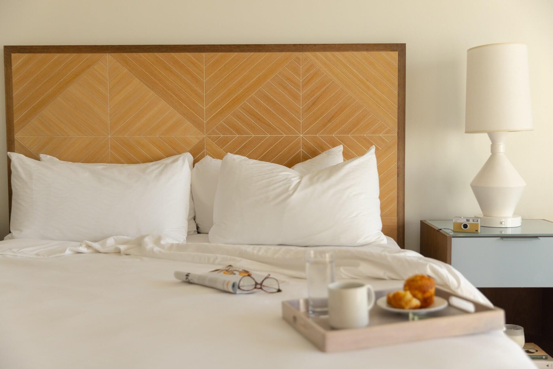 Cozy hotel bed with wooden headboard, white linens, bedside lamp, and breakfast tray with coffee and pastries.
