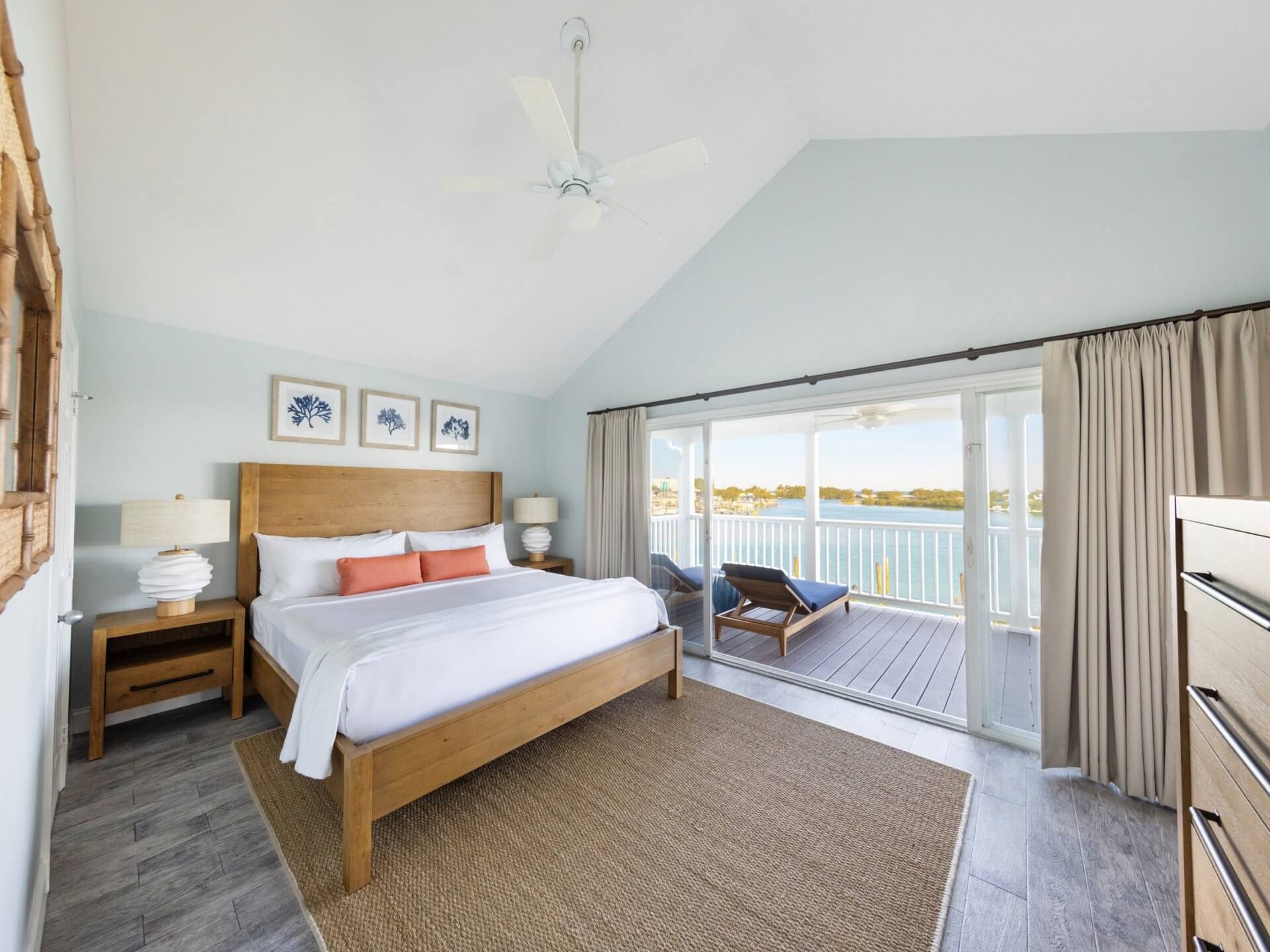 Waterfront resort bedroom with king bed, vaulted ceiling, and sliding doors opening to a balcony with lounge chairs.