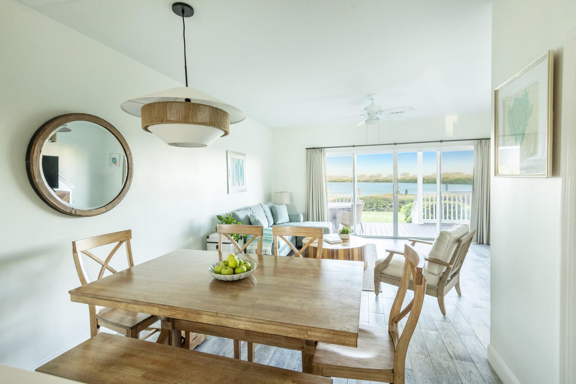 Bright coastal dining and living area with wooden table, seating, and sliding glass doors opening to a water view.
