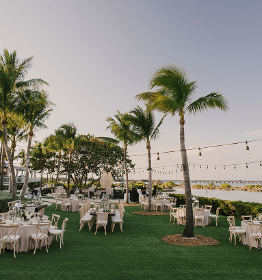 Outdoor waterfront event with round tables on a lawn, palm trees, and string lights overlooking calm water at sunset.