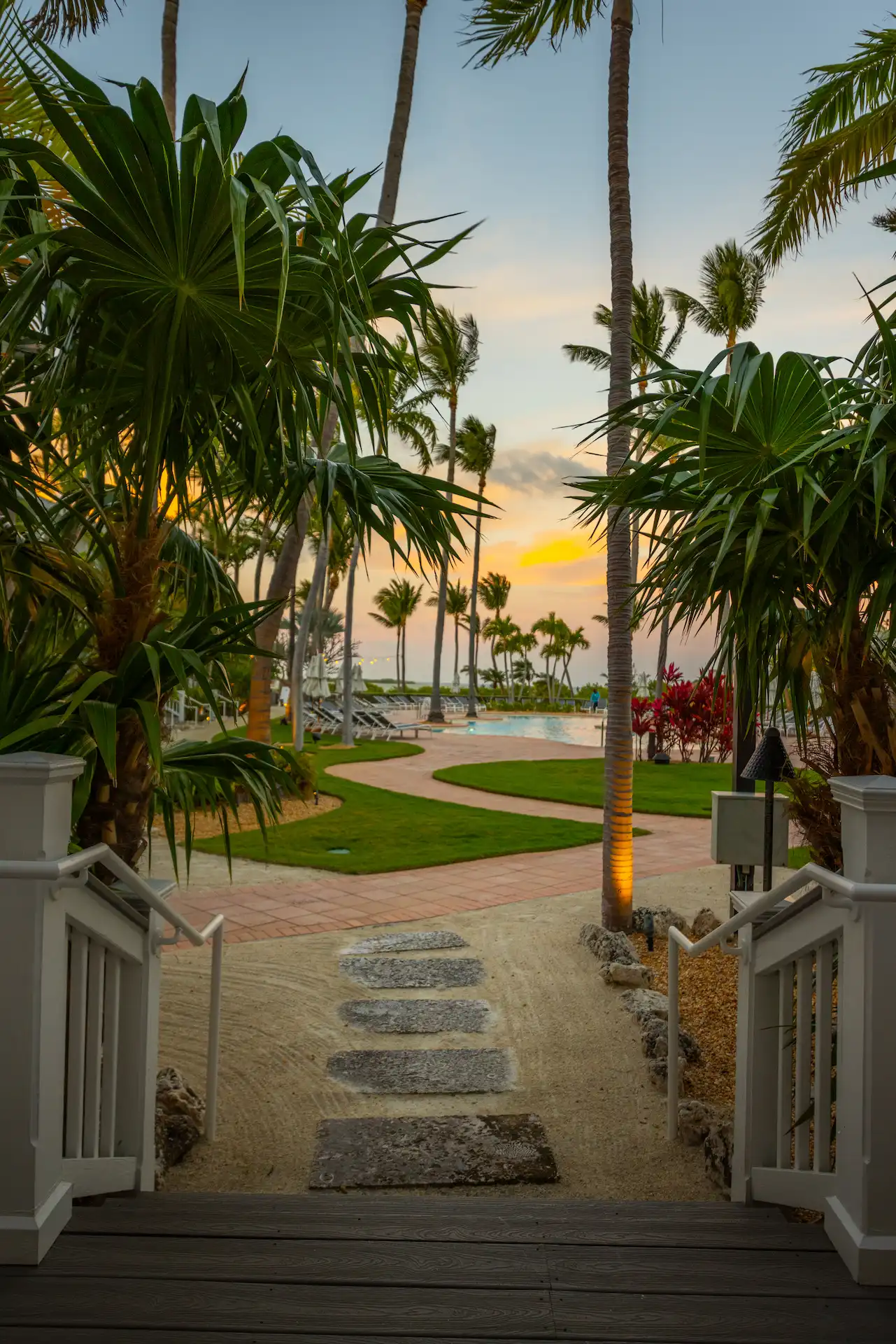 Palm-lined resort walkway leading to a pool at sunset, with tropical landscaping and soft evening light.