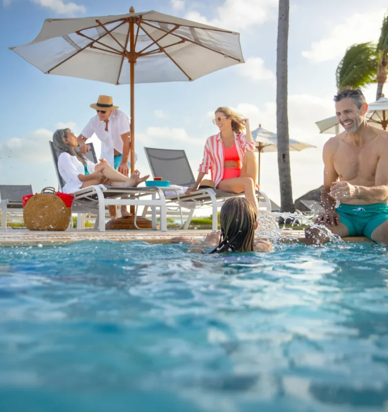 Family relaxing and playing in a resort swimming pool with lounge chairs, umbrellas, and palm trees on a sunny day.