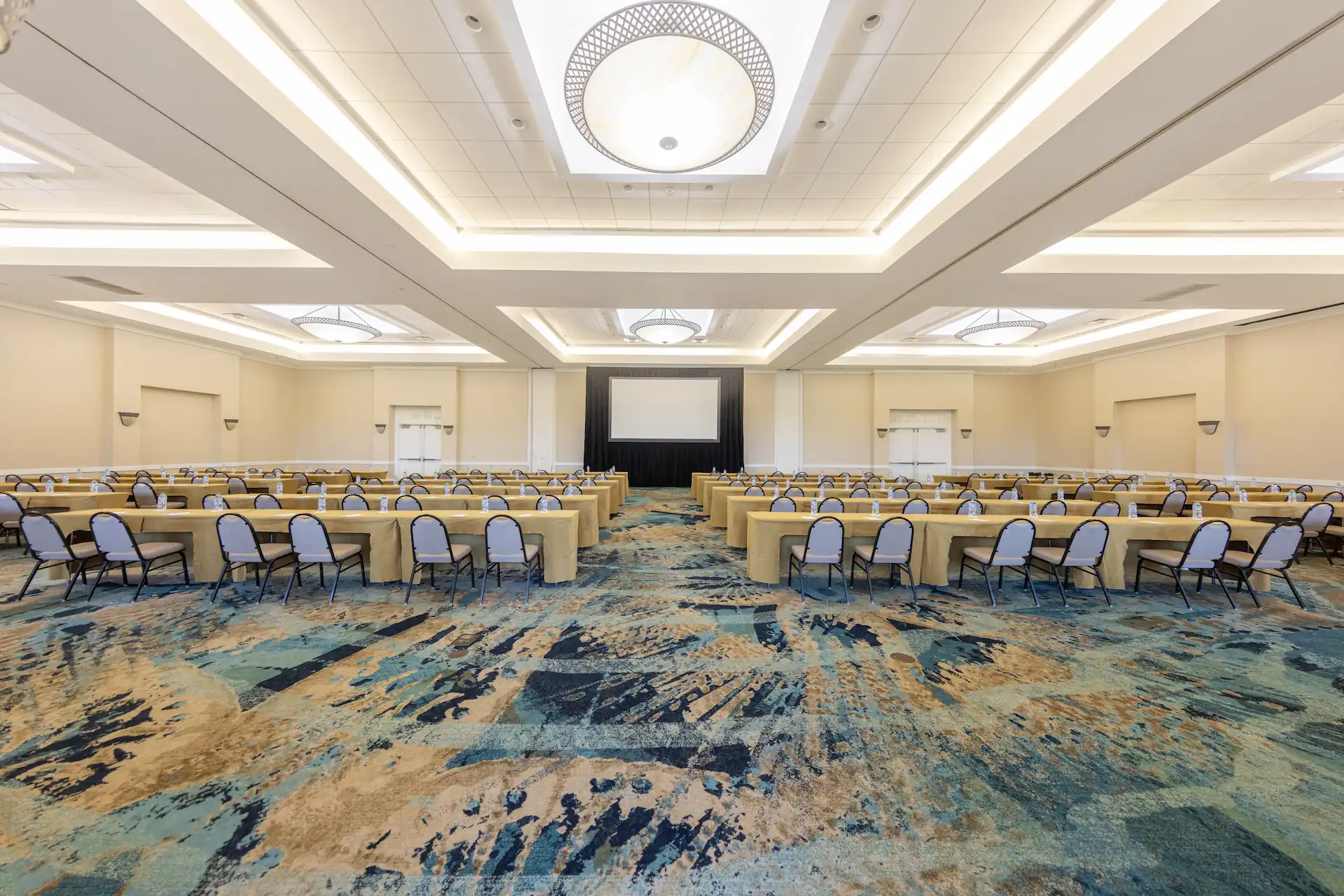 Large conference ballroom with classroom-style tables, chairs, projection screen, and modern ceiling lighting.