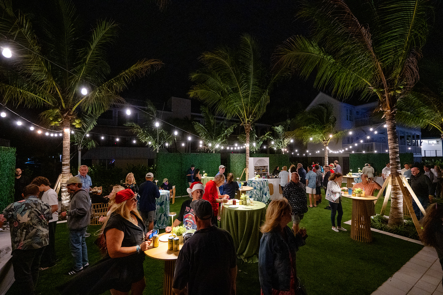Nighttime outdoor gathering with guests mingling under string lights and palm trees in a tropical courtyard.