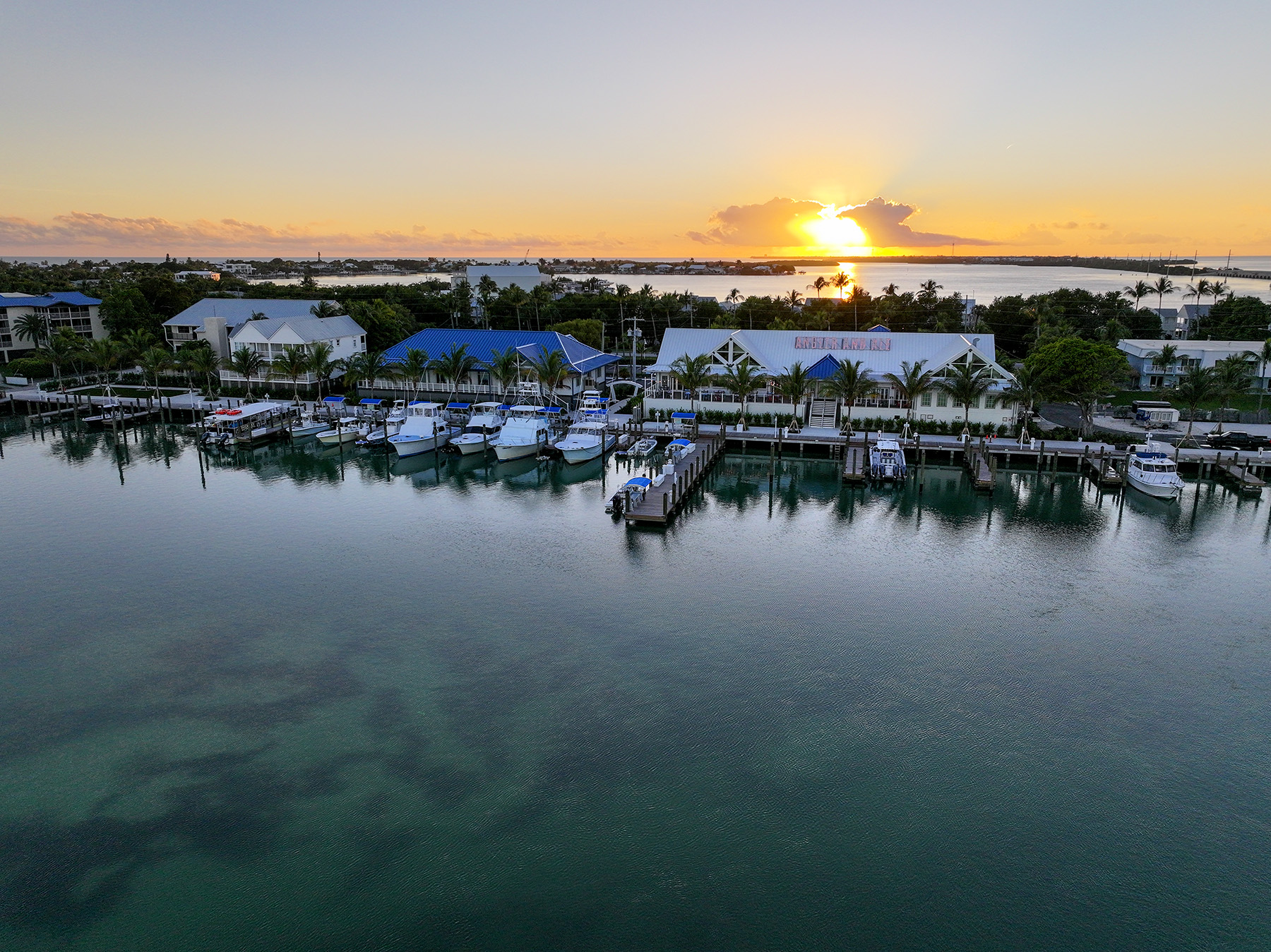Aerial view of a marina at sunset with docked boats, palm trees, and waterfront buildings reflected in calm water.