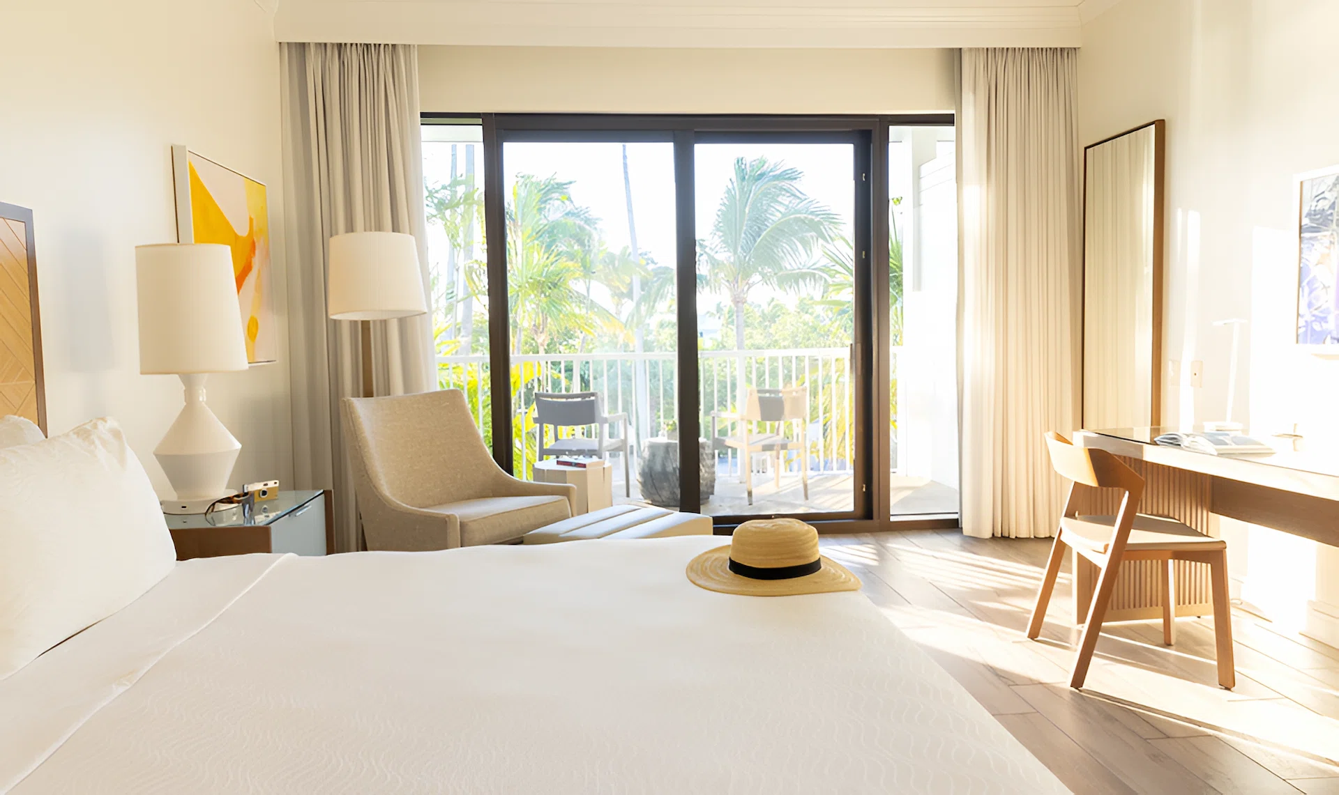 Sunlit coastal bedroom with white bedding, desk and chair, and sliding glass doors opening to a balcony with palm views.