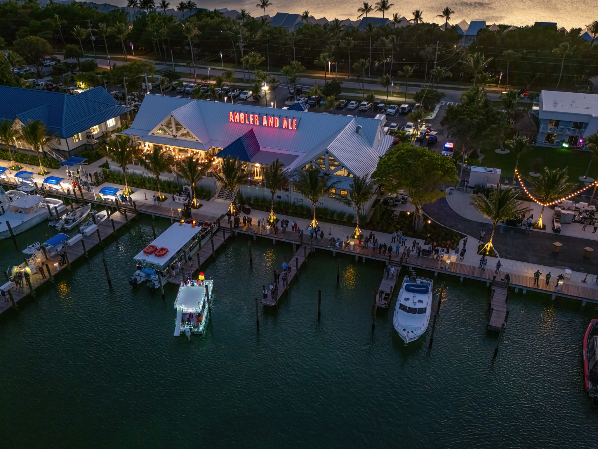 Aerial dusk view of Angler and Ale waterfront restaurant with marina docks, boats, palm trees, and glowing lights.