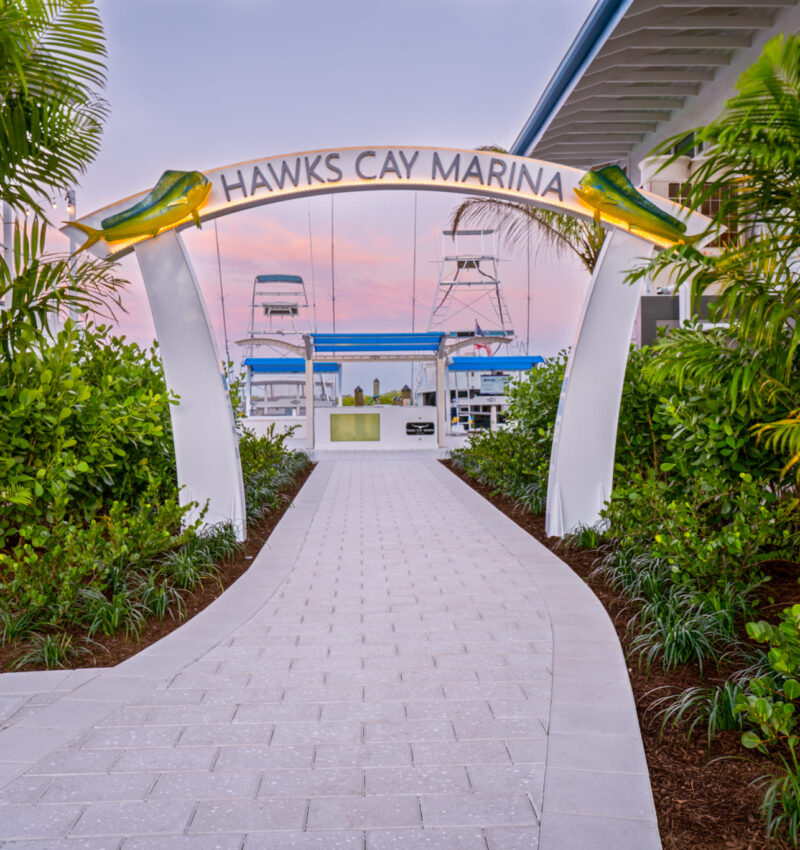 Entrance to Hawks Cay Marina with palm-lined walkway, fish sculptures on the arch, and boats visible beyond.