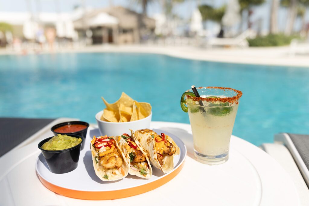 Plate of fish tacos with chips, guacamole, salsa, and a margarita served poolside at a sunny resort.