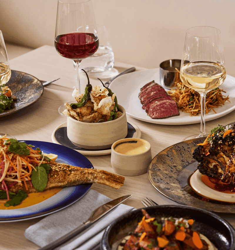 Table set with gourmet dishes and wine glasses, featuring steak, fish, vegetables, and small plates in an upscale restaurant.