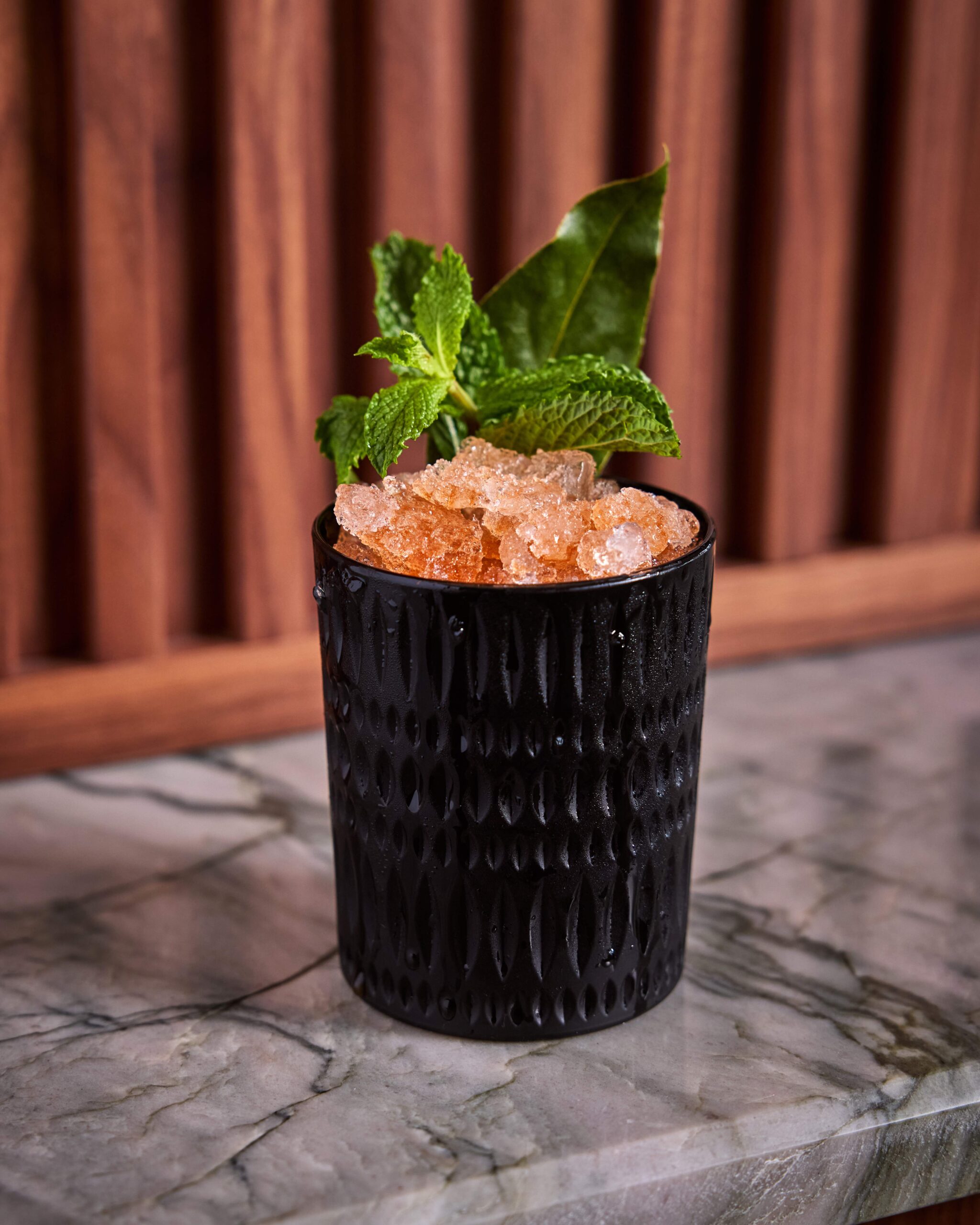 Black cocktail glass with crushed ice, garnished with fresh mint leaves on a marble surface.