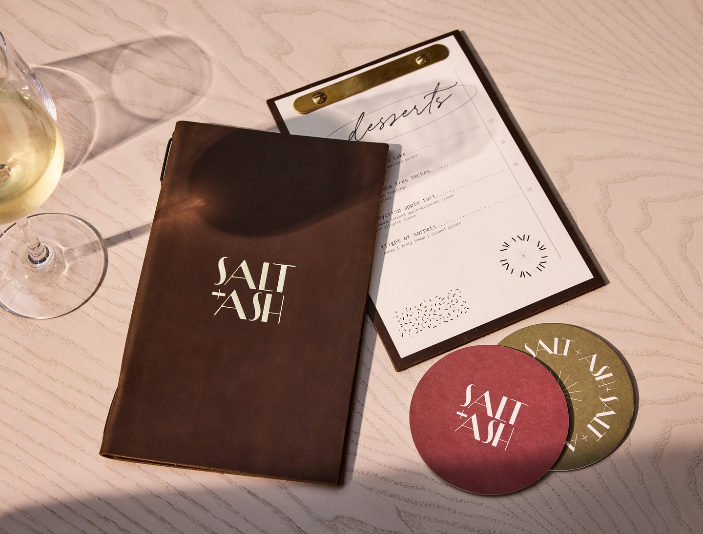 Salt + Ash restaurant menu and dessert card on a table with branded coasters and a glass of white wine.