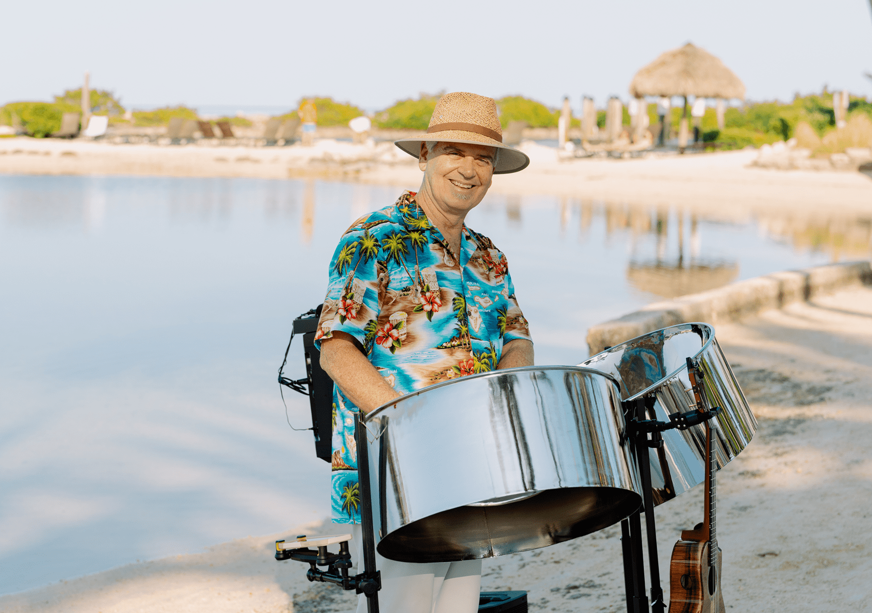 Smiling steel drum musician in a tropical shirt performing by a calm lagoon with beach chairs and palm trees nearby.