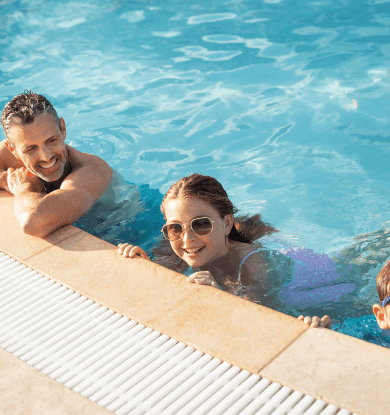 Smiling adult and two children wearing sunglasses relax at the pool edge in clear blue water on a sunny day.