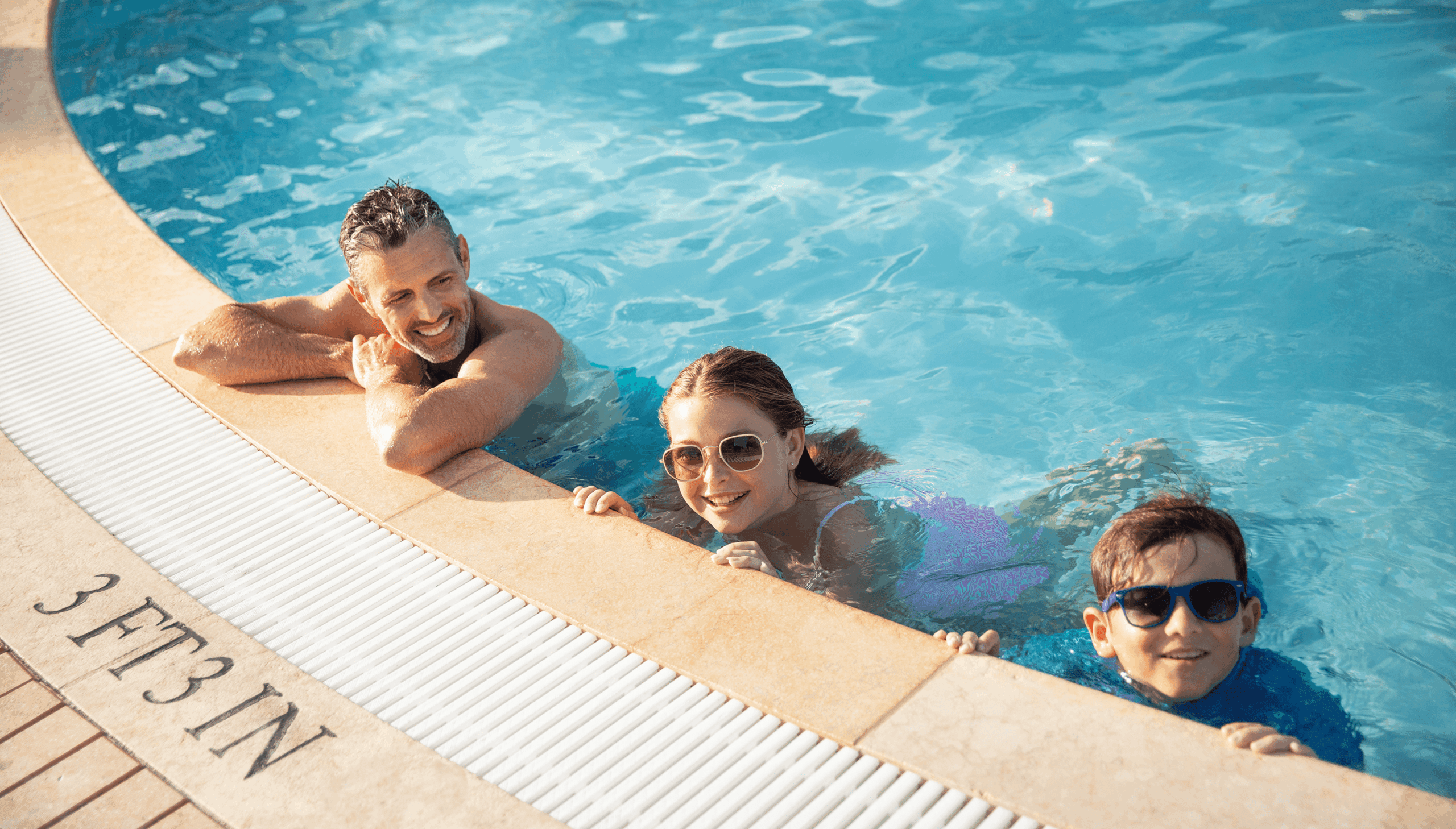 Smiling adult and two children wearing sunglasses relax at the pool edge in clear blue water on a sunny day.